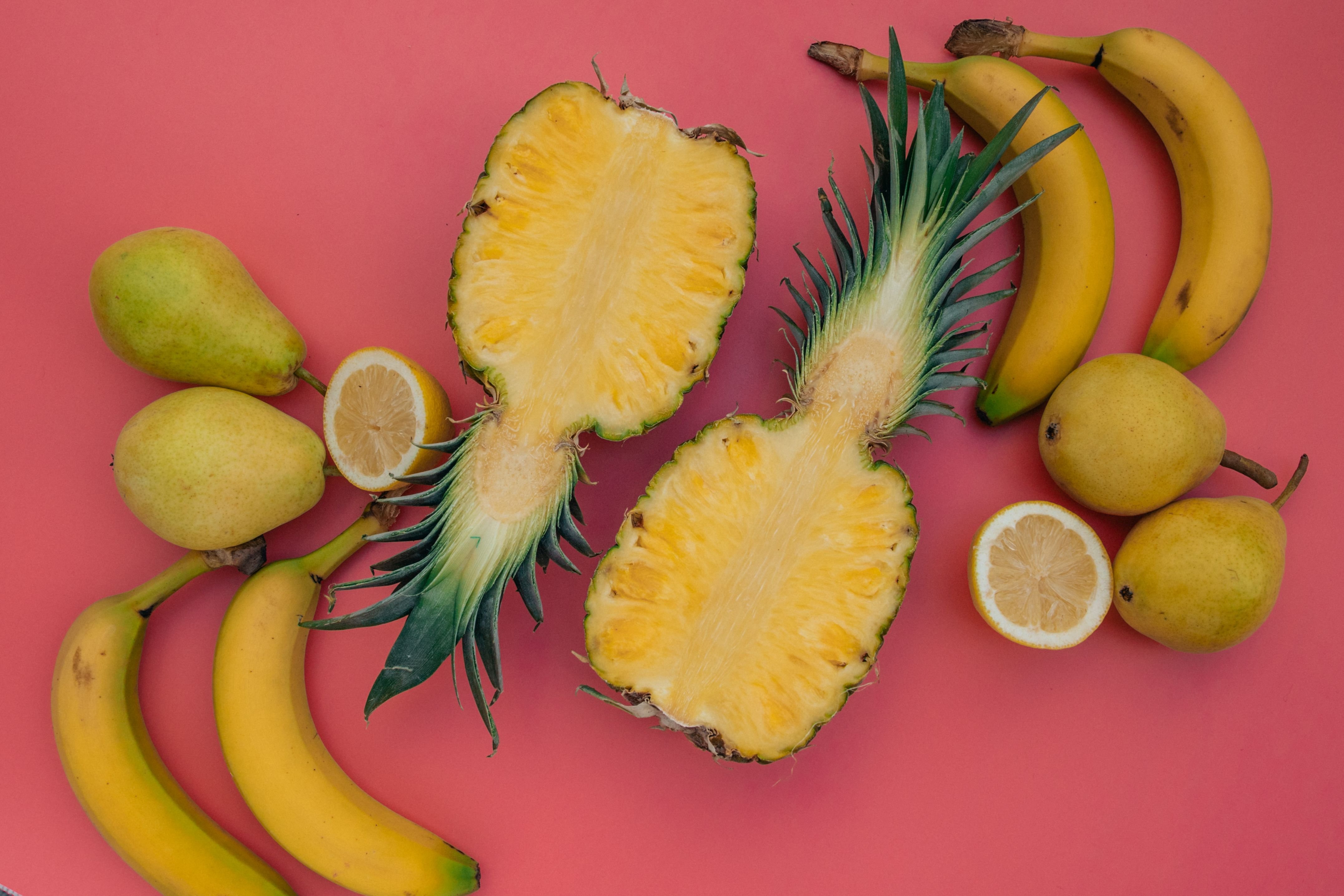 Vibrant still life of sliced pineapple, bananas, pears, and lemons on a pink background.